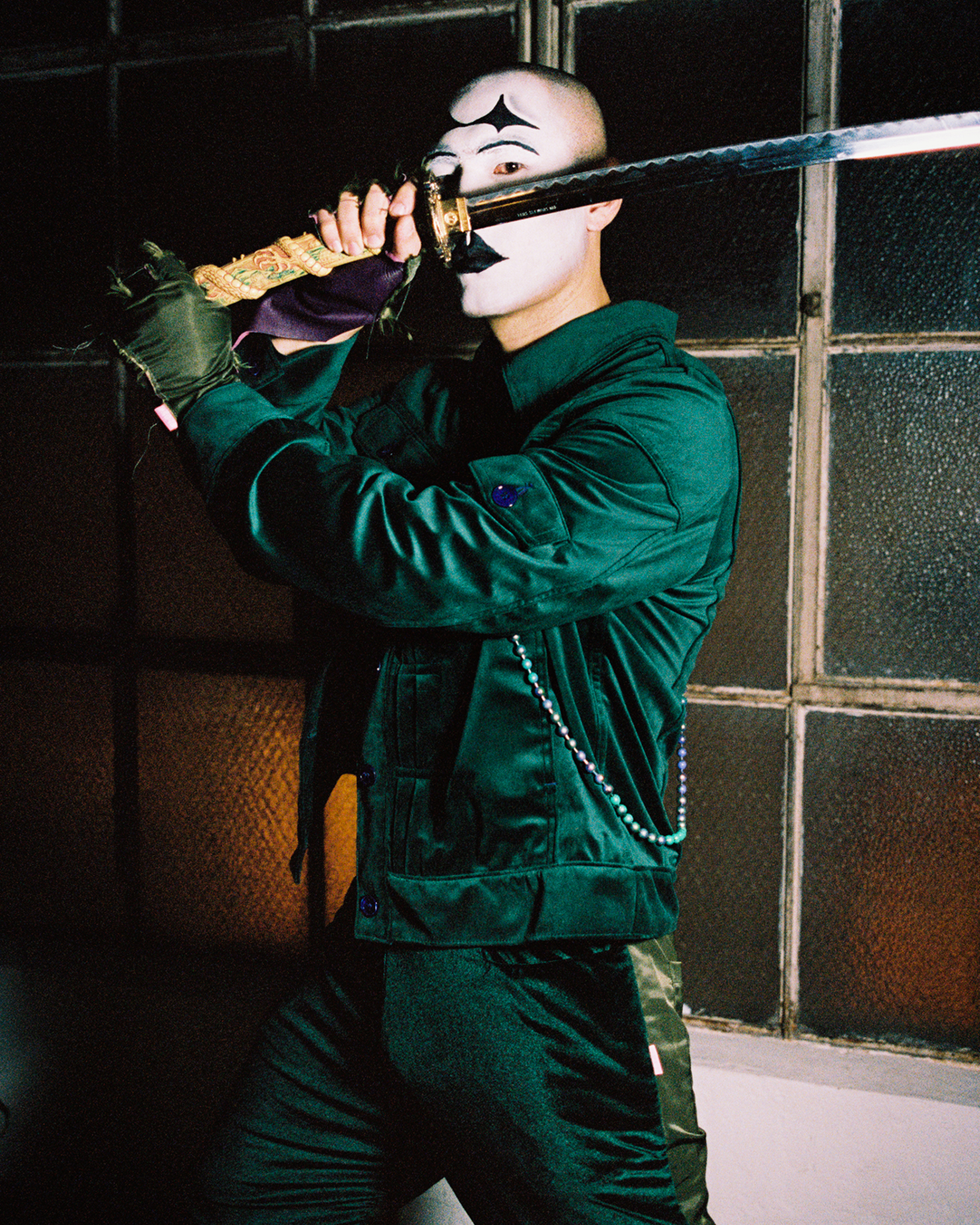 Model with black and white joker facepaint wearing a green ensemble wealding a sword in fron of his face